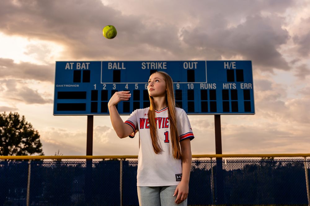 A softball soars through a dramatic cloudy sky at sunset during girl senior photos in Omaha, NE. - - by BrightSide Photography