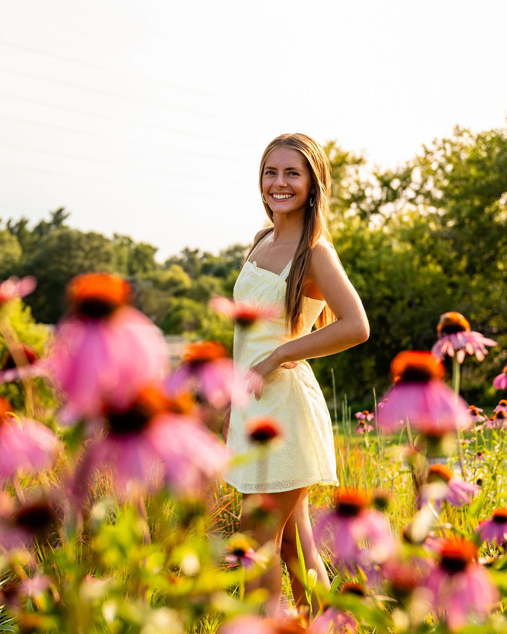 A senior girl in a yellow dress poses joyfully among vibrant pink coneflowers in a summer garden at golden hour. - by BrightSide Photography