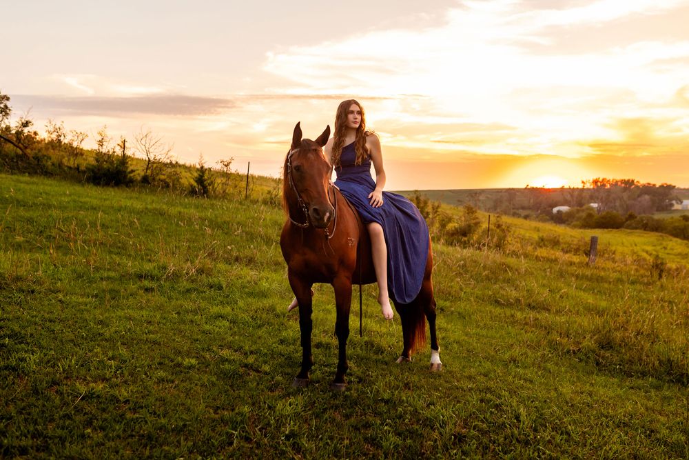 A horse and senior girl stand together in a grassy field at sunset creating a silhouette against the golden sky. - by BrightSide Photography