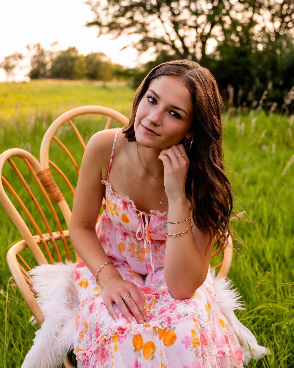 A person in a floral pink sundress poses on a wooden chair in a sunlit meadow at golden hour.