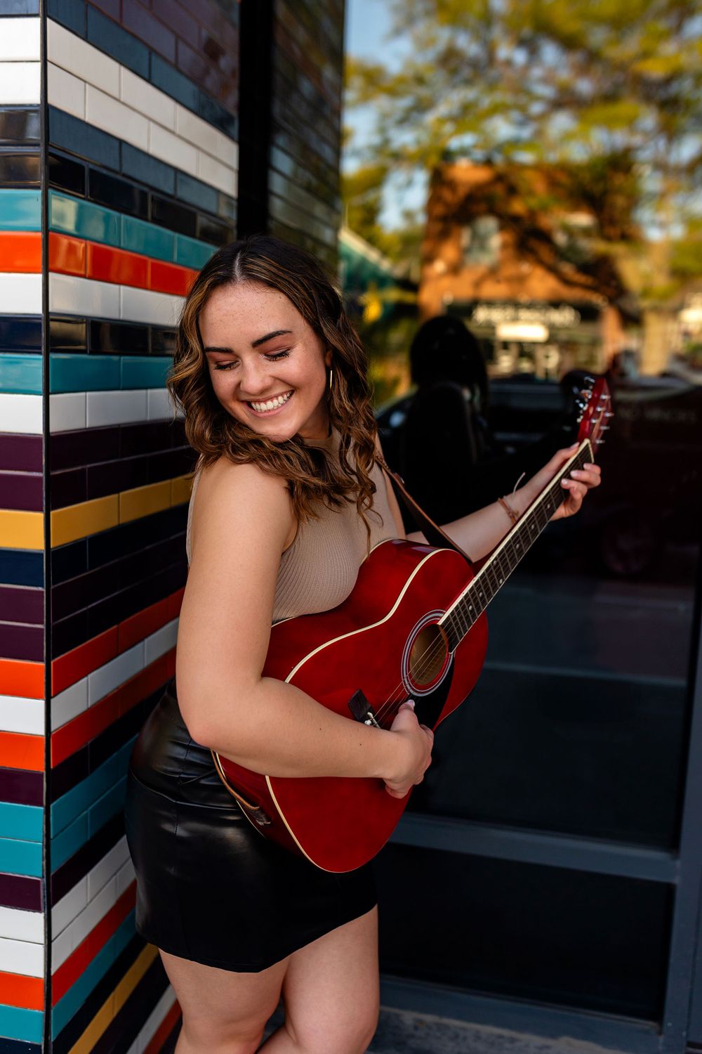 A senior girl musician poses with acoustic guitar against colorful striped wall in outdoor portrait session. - by BrightSide Photography
