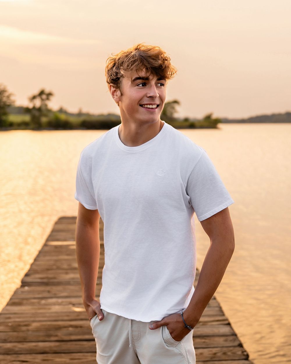 Person in white t-shirt poses on wooden dock during golden sunset at lake.