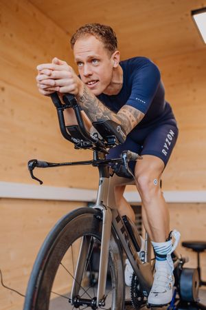 Indoor cyclist wearing navy blue athletic gear trains on a black stationary bike against a wooden wall background.