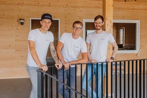 Three people in white t-shirts and jeans smile while standing together on a wooden porch with black railings.