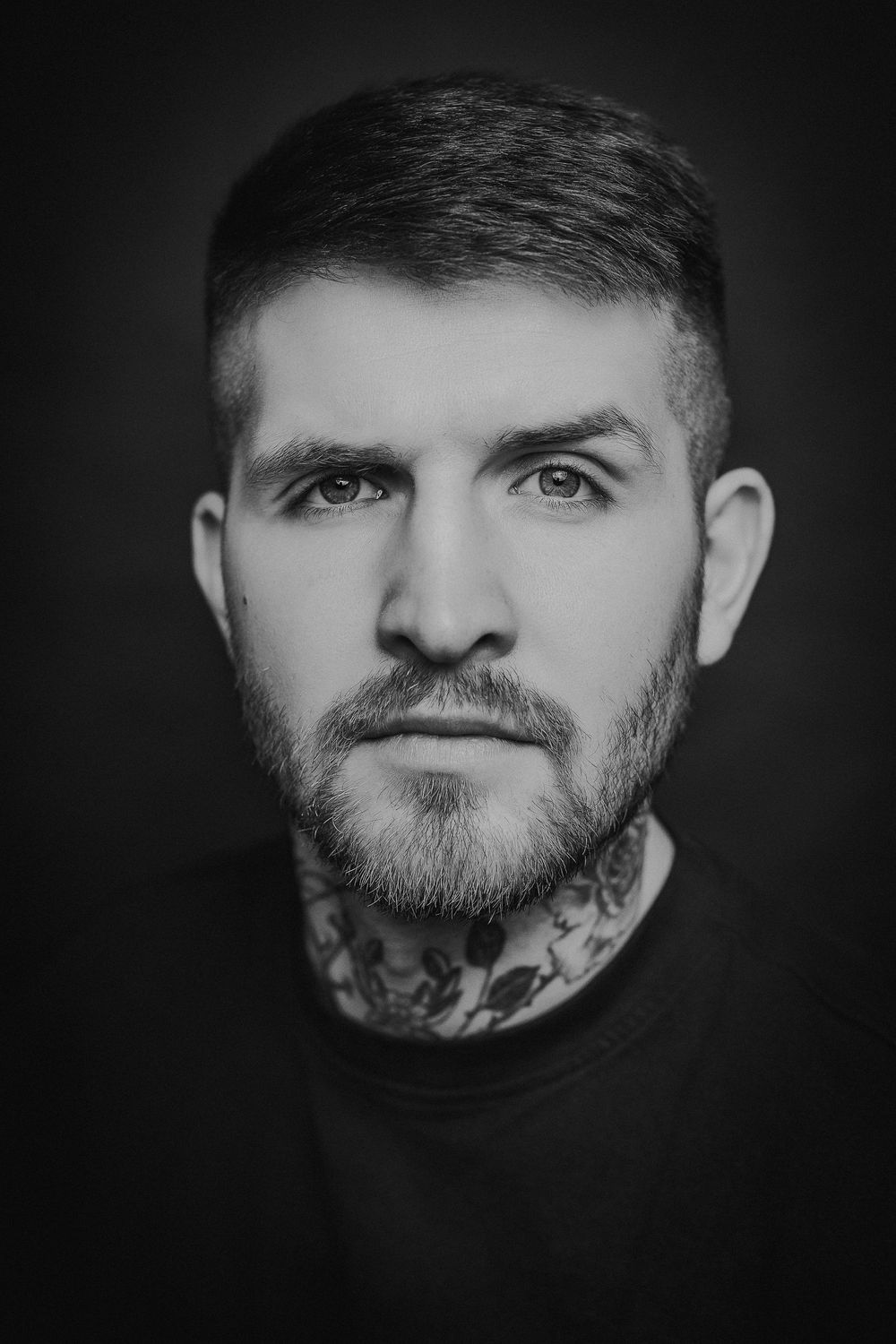 Dramatic black and white portrait showing neck tattoos and dark clothing against a black background.