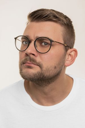 Portrait of a contemplative person wearing round glasses and a white t-shirt against a light background.