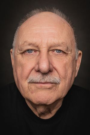 Close-up portrait of a senior man with a mustache wearing dark clothing against a dark background.