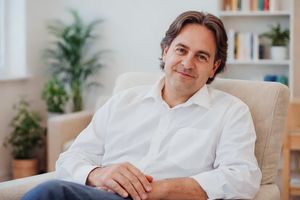 Professional in a white button-down shirt relaxes on a modern gray couch in a bright home office setting.