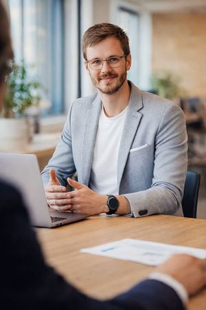 Professional in a gray blazer and white shirt sitting at wooden desk in modern office setting.