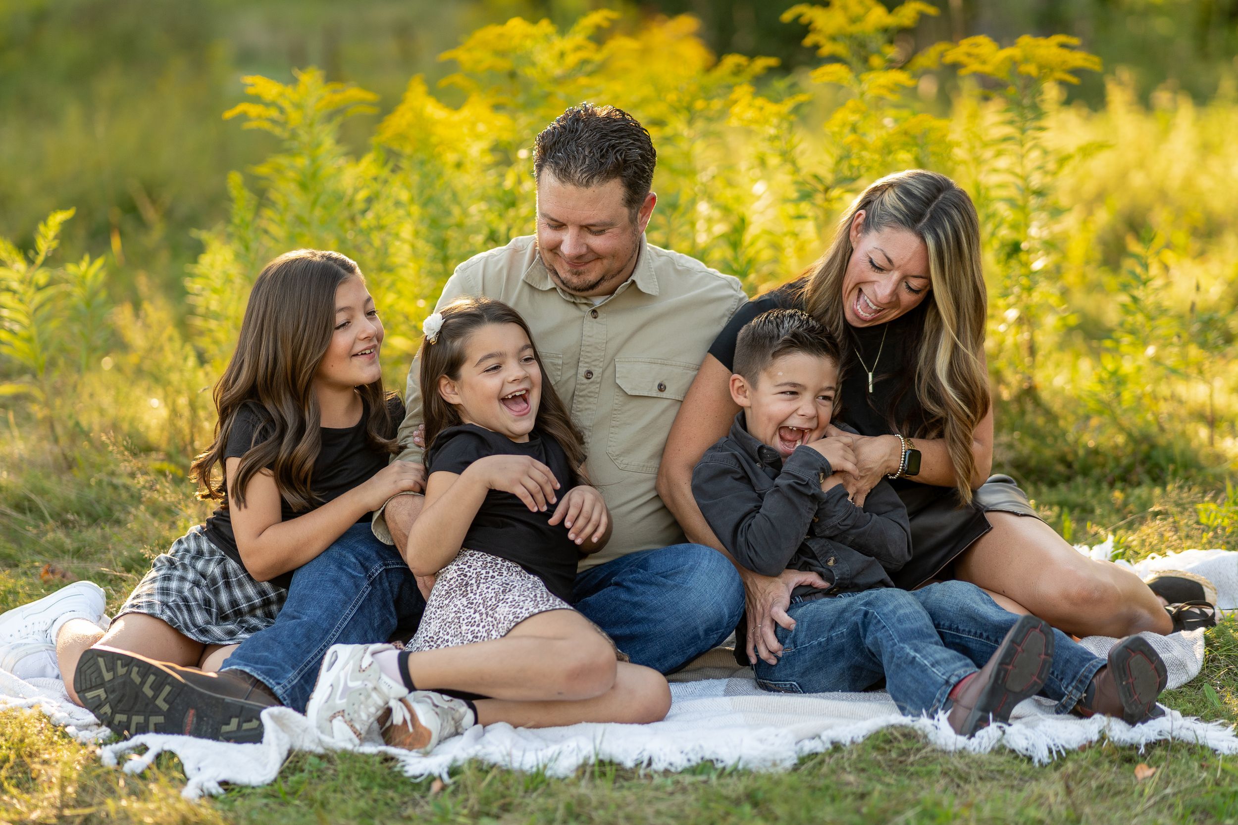 A family shares joyful laughter while sitting together on a white blanket surrounded by golden wildflowers in nature.