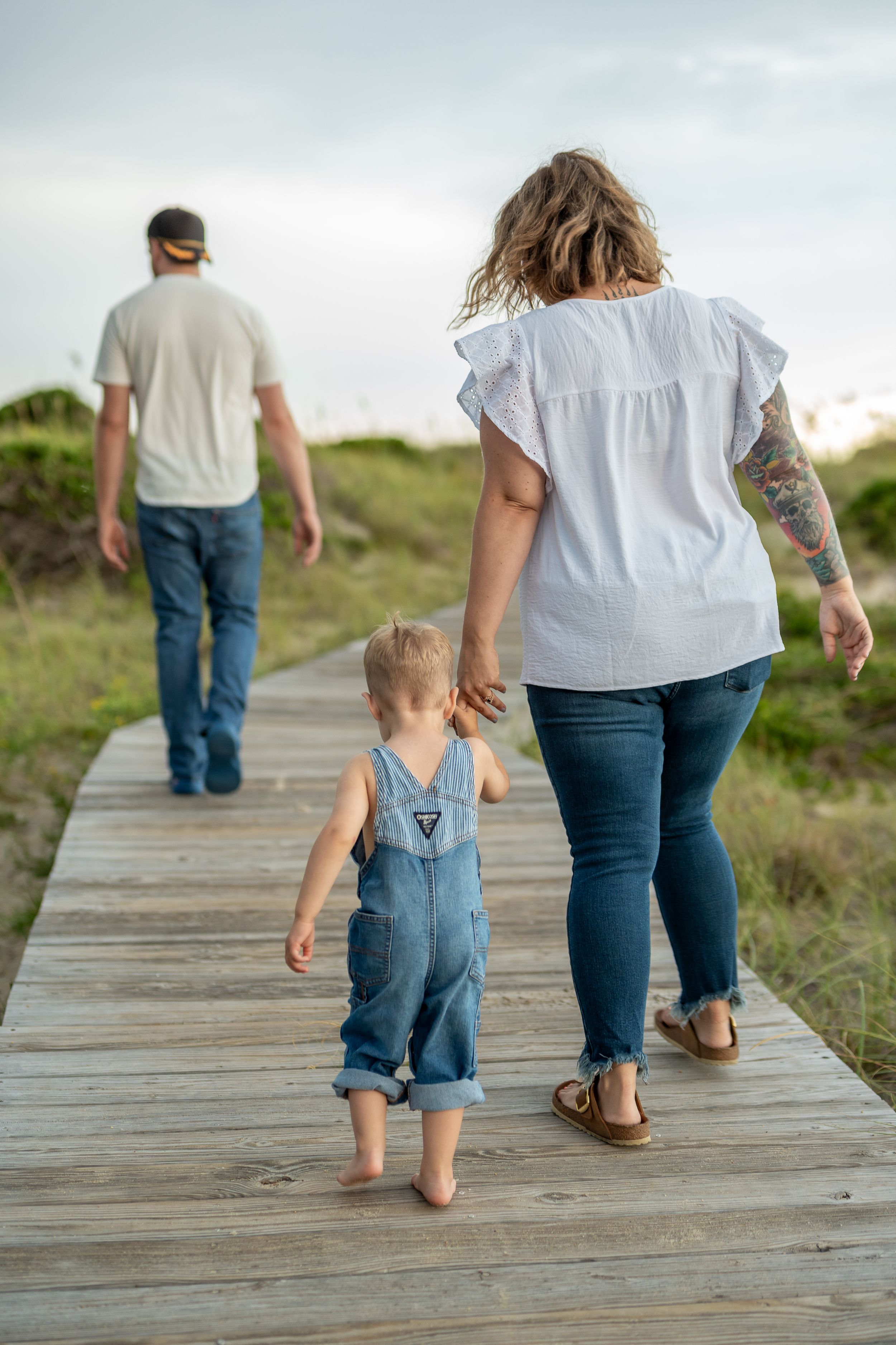 A family walking barefoot on a wooden boardwalk path through natural scenery.
