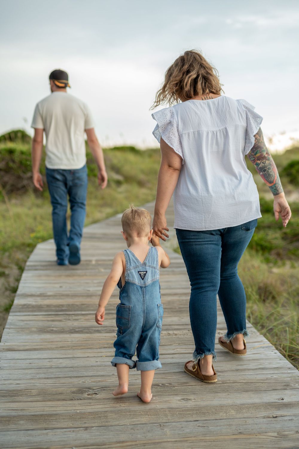 A family walking barefoot on a wooden boardwalk path through natural scenery.