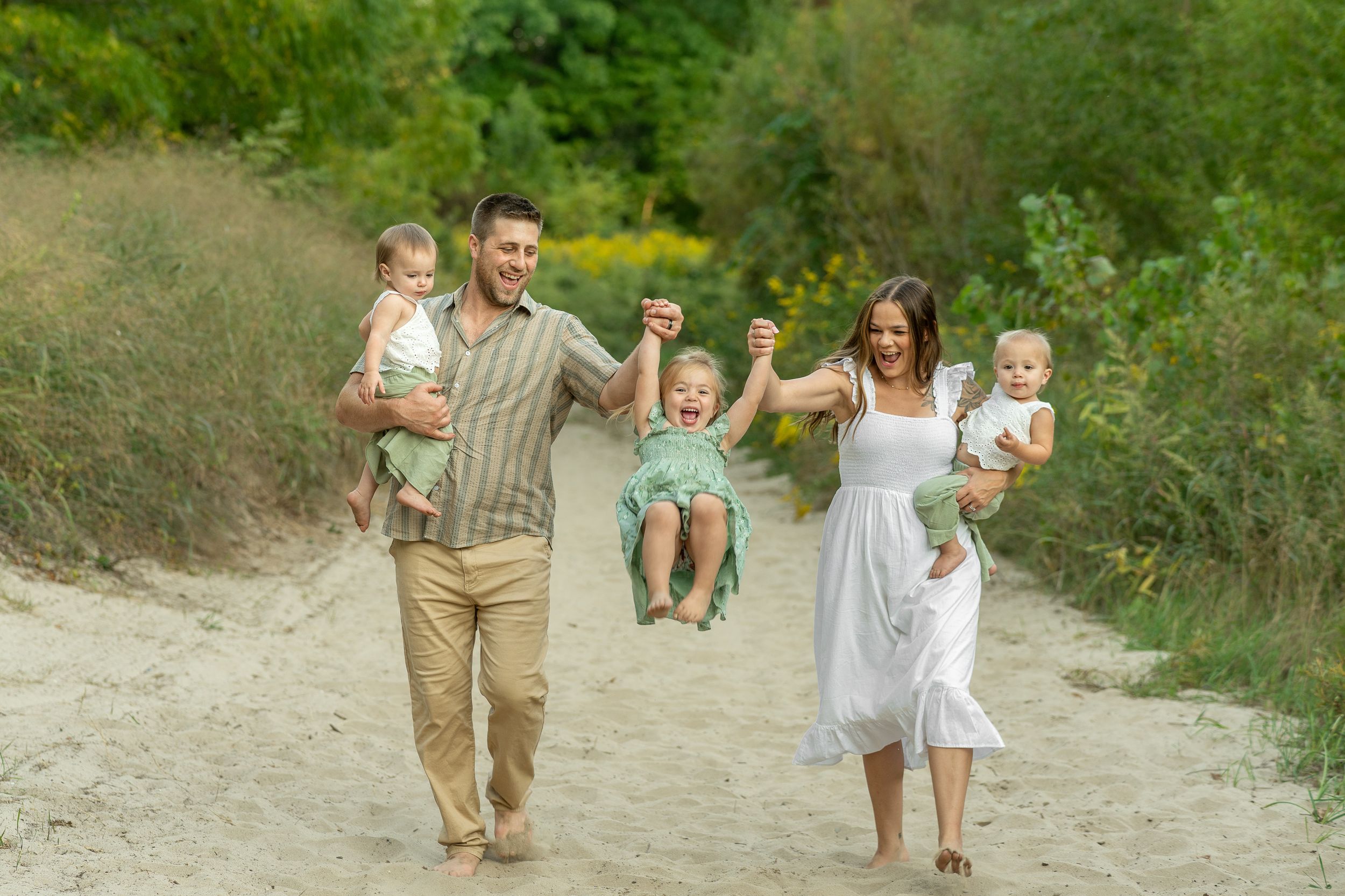 A group of people playfully chase each other down a sandy beach path surrounded by green foliage.