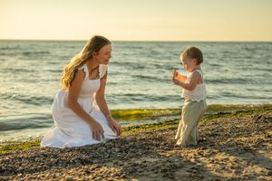 A mother and child share a tender moment while playing on a sandy beach at sunset by the ocean.