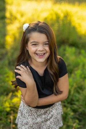 A young girl in a black top and patterned skirt poses playfully in a sunny outdoor setting with yellow flowers.