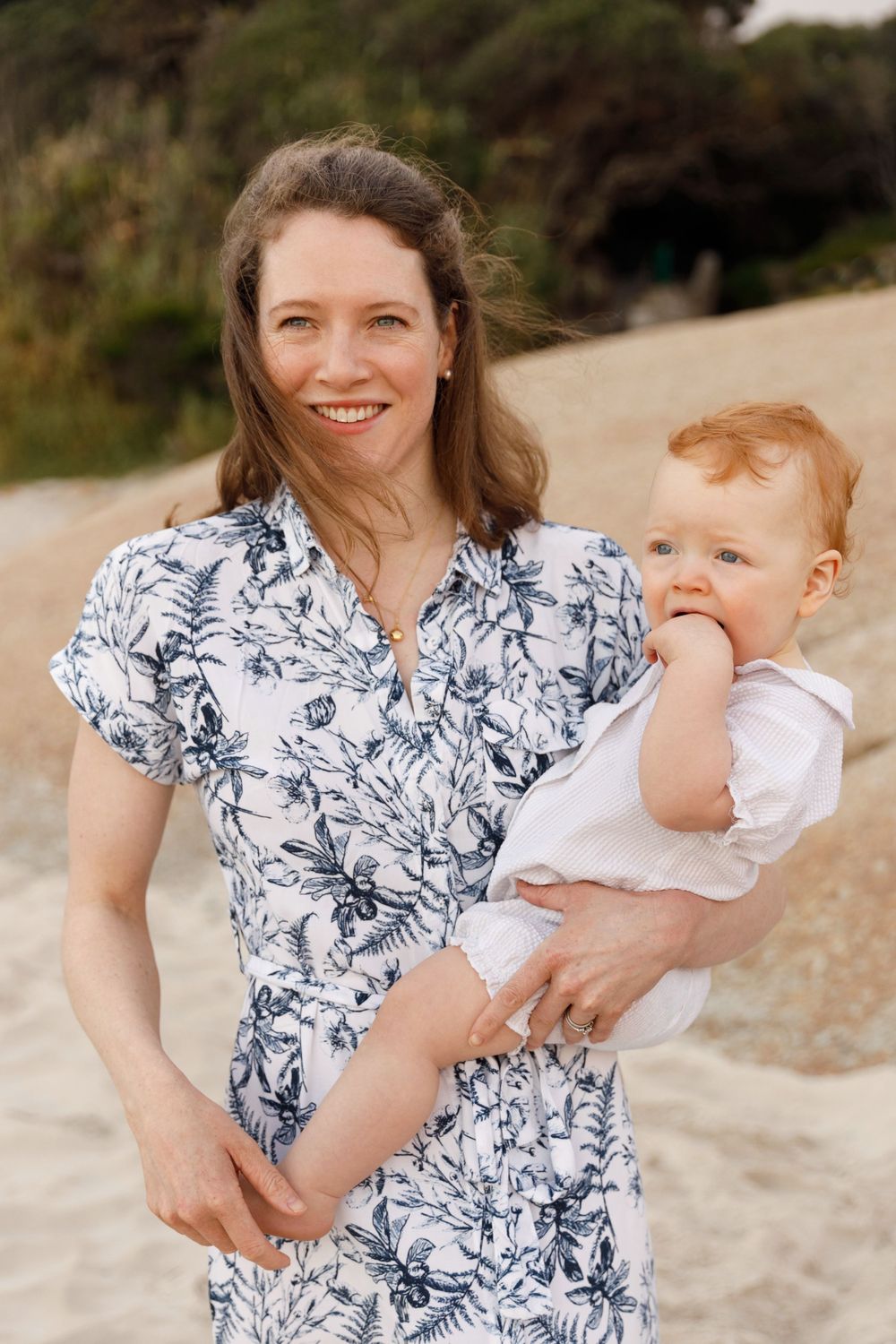 A woman wearing a blue and white floral dress holds a baby in white clothing on a sandy path outdoors.