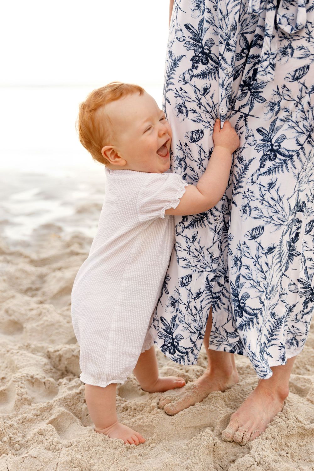 Laughing baby in white romper holds onto floral dress while standing on sandy beach.