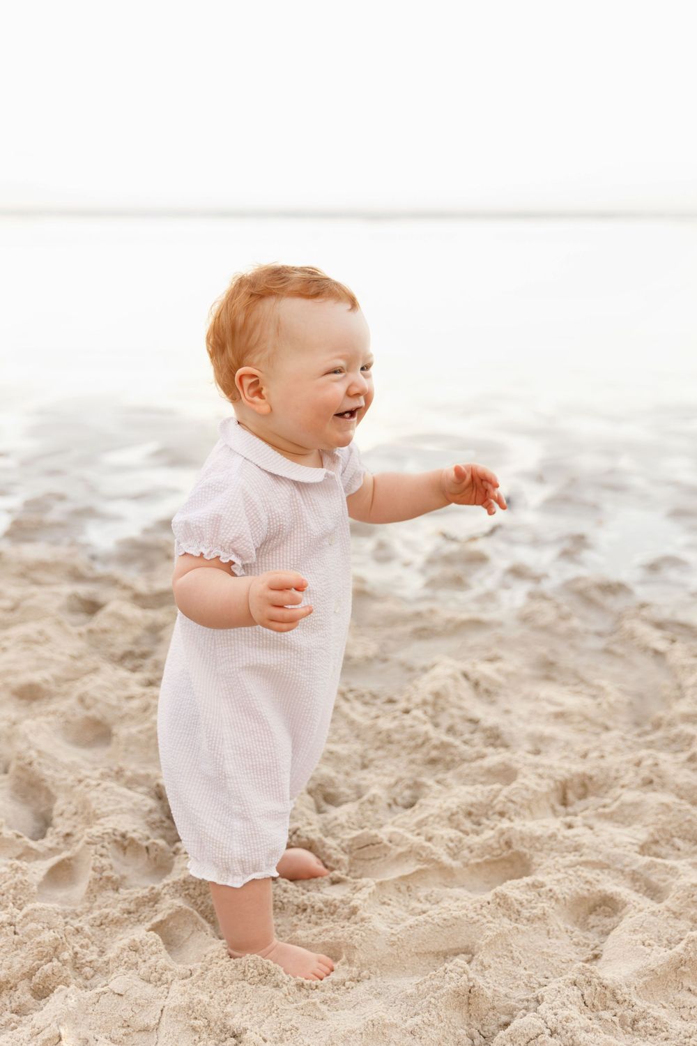 Laughing baby in white romper standing barefoot on sandy beach in soft lighting.
