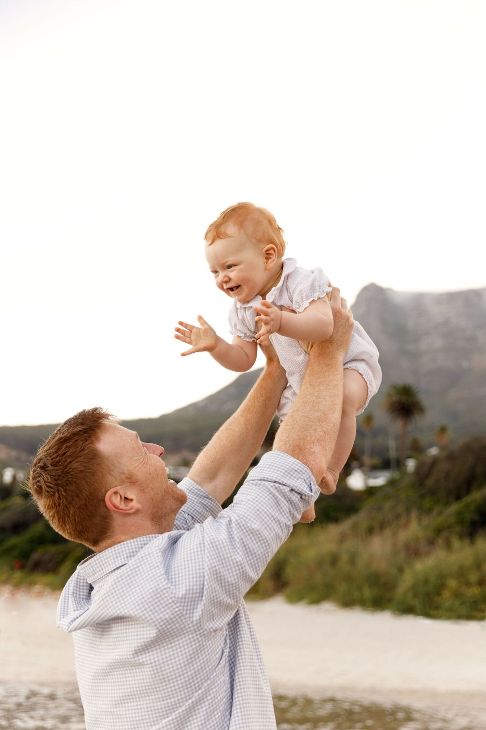 Parent lifting joyful baby against backdrop of mountain beach landscape.