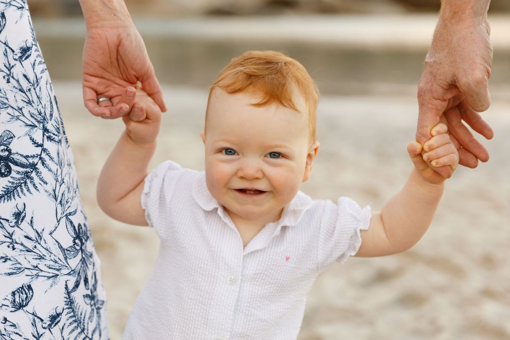 A small child in a white shirt smiles while holding hands and walking on the beach.
