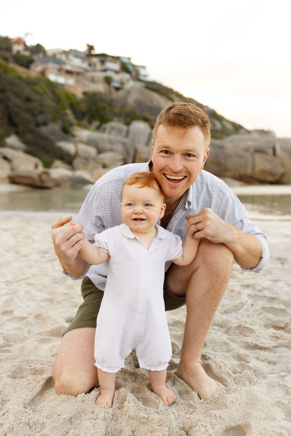 A joyful moment between parent and toddler standing on a sandy beach with coastal homes in the background.