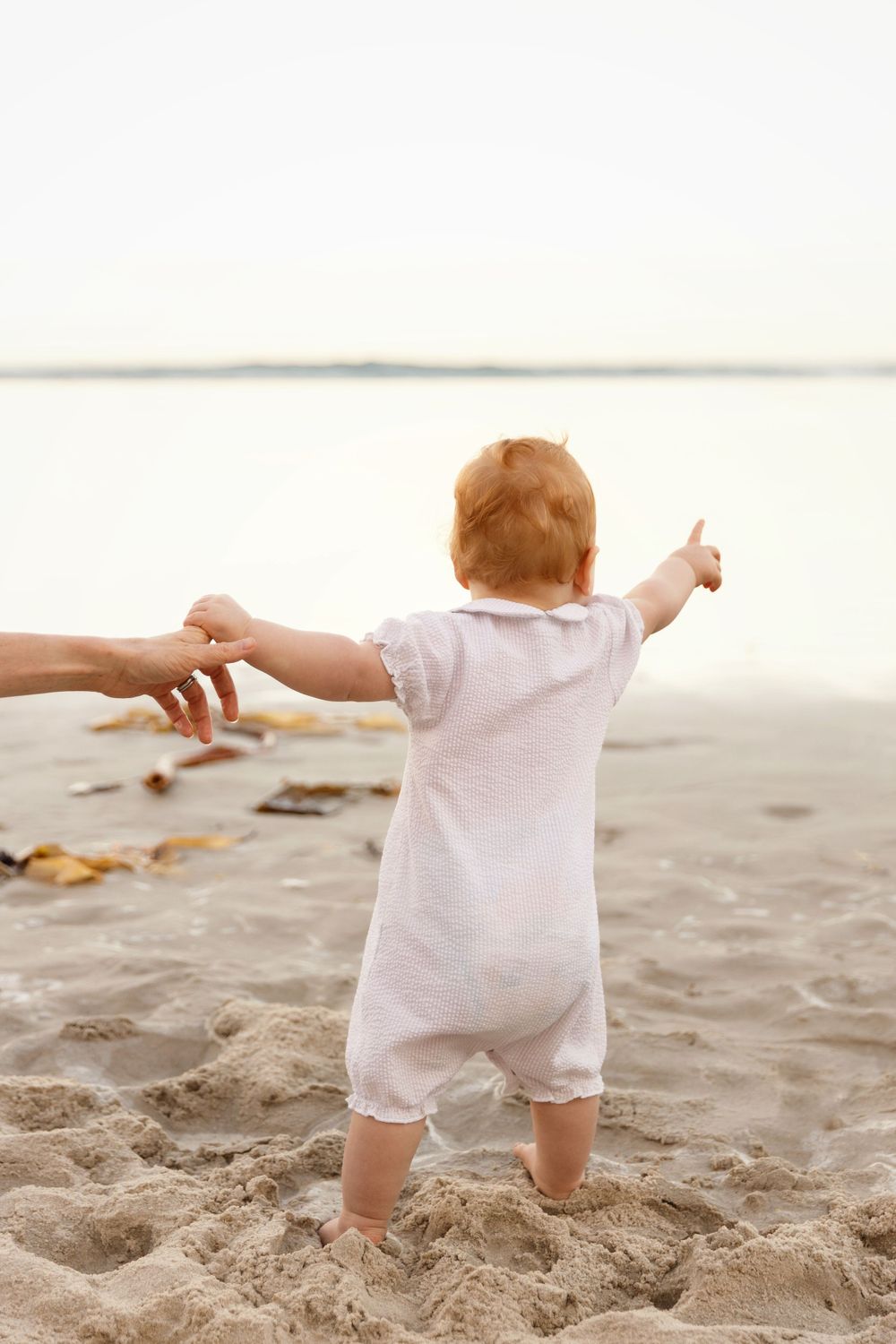 Baby in white outfit learning to walk on beach while holding adult's hands.