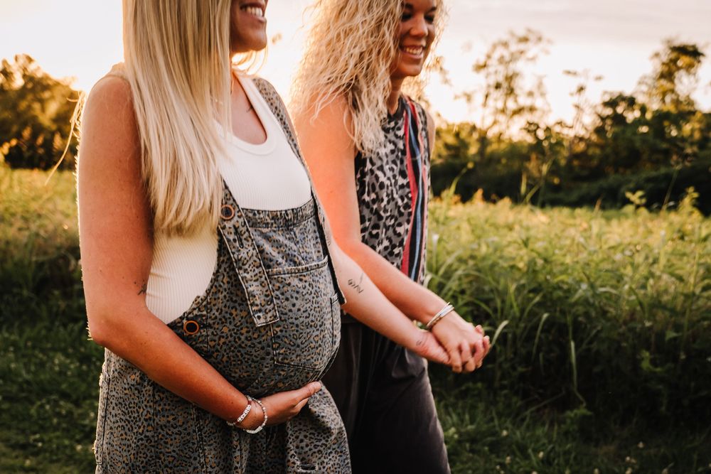 Two women mothers standing outdoors at sunset, one visibly pregnant  in a green field. same sex maternity photoshoot nashville, tennessee
