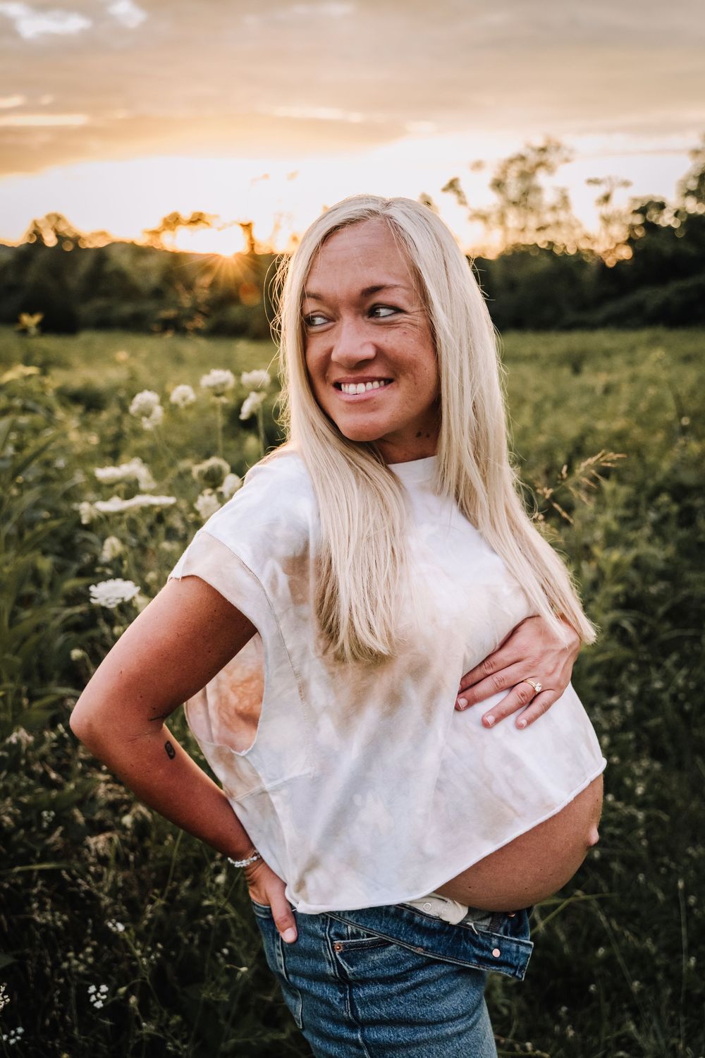 Smiling pregnant woman with blonde hair posing in a flower field at golden sunset, wearing white tee and jeans. nashville tn maternity photo