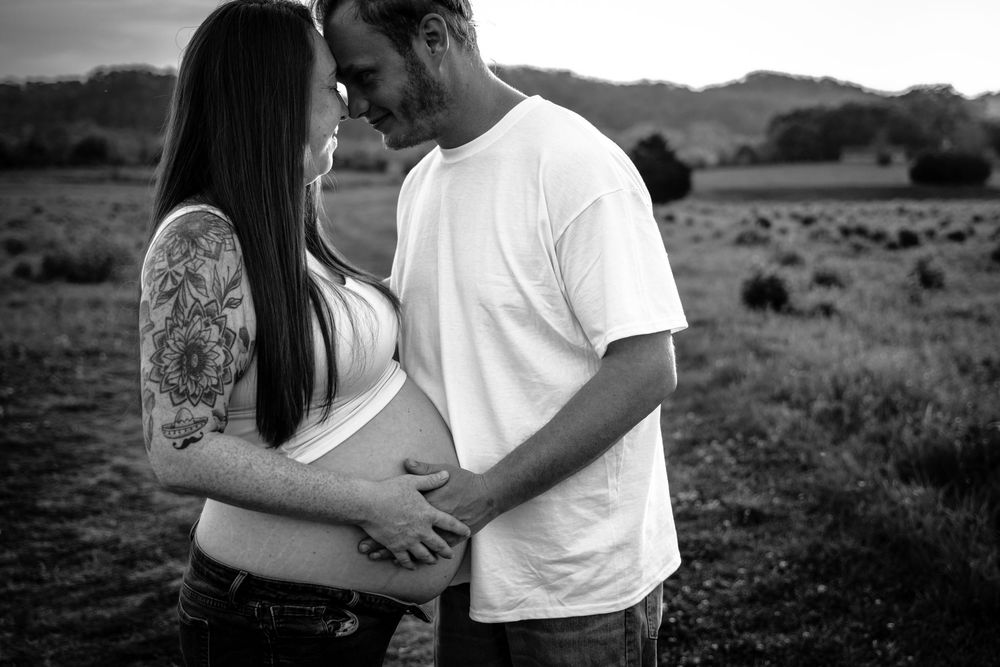 Black and white photo of a pregnant couple holding baby bump in a field, sharing a tender moment outdoors. nashville pregnancy photographer