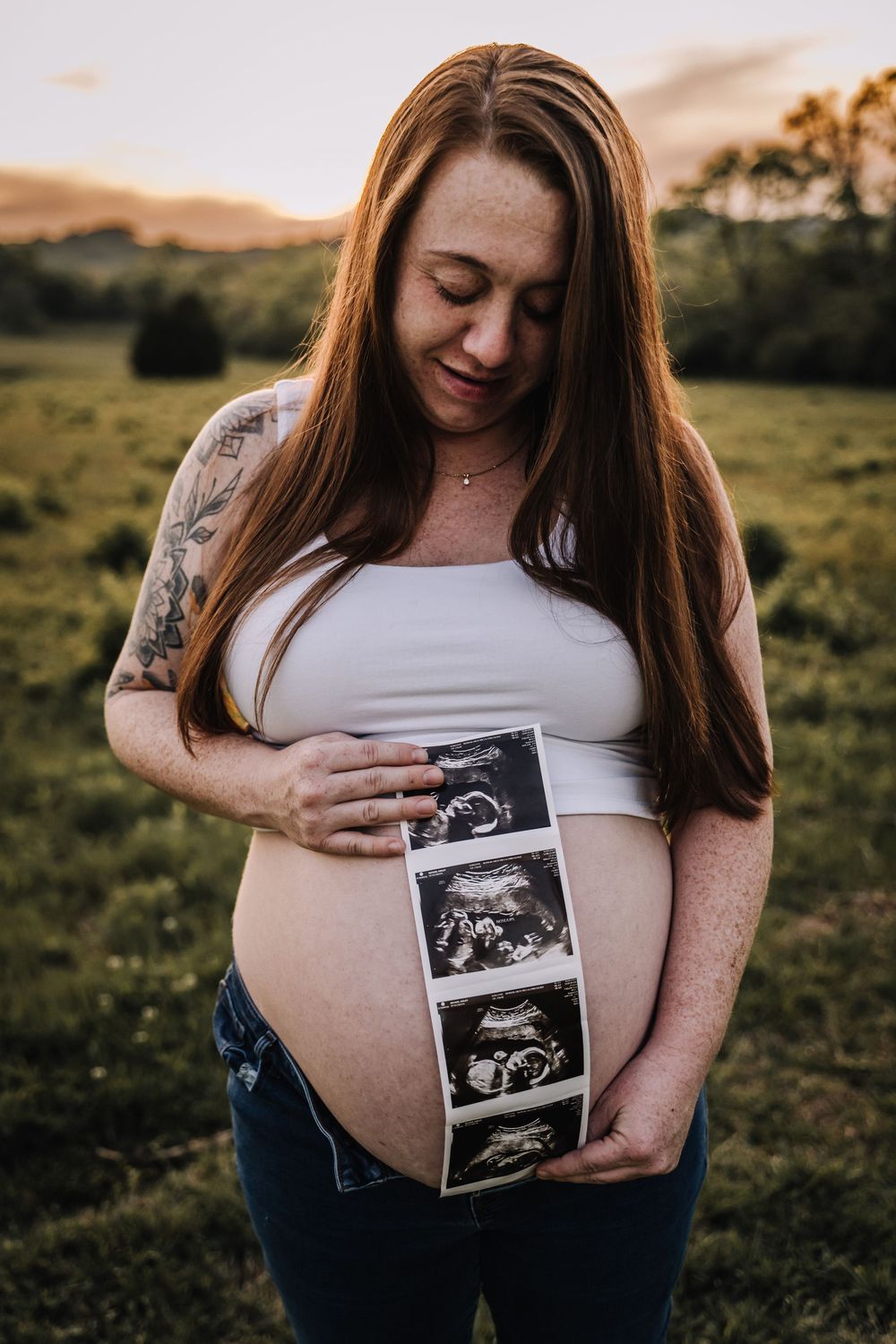 Pregnant woman holding ultrasound photos against her baby bump outdoors at sunset, wearing white crop top and jeans. middle Tennessee photog