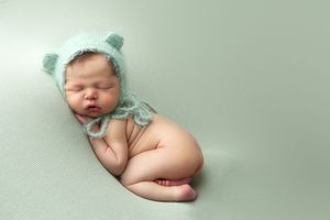 Sleeping newborn wearing soft mint green bear hat lies curled up on matching mint backdrop during professional photo session.