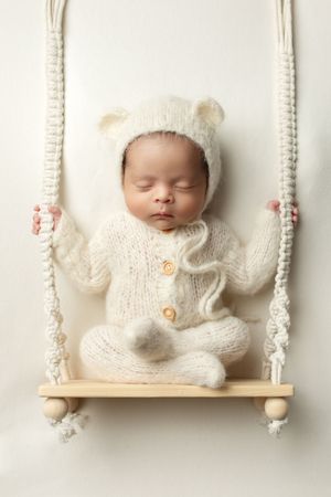 Sleeping baby wearing cozy white knit outfit with bear ears rests peacefully on decorative wooden swing.