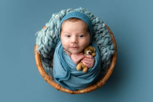 Newborn baby wrapped in soft blue blankets lies peacefully in a round wooden basket against blue backdrop.