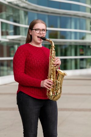 A musician wearing glasses and a red sweater poses with a saxophone in front of a modern glass building.