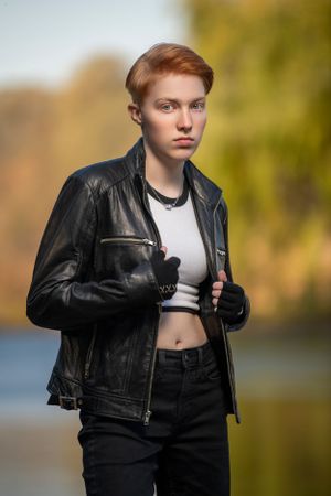 A person with a cropped white top and black leather jacket poses outdoors against a blurred autumn background.
