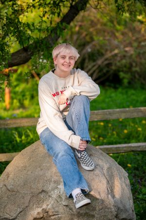 Someone sits casually on a large rock wearing a cream sweater, jeans and checkered Vans sneakers in a sunny garden setting.