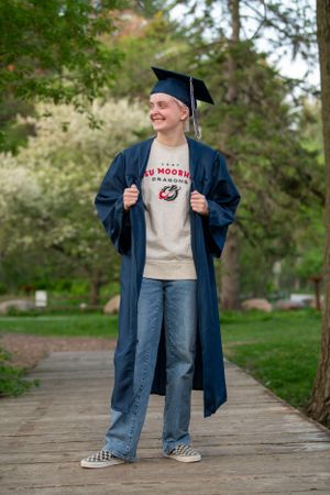 Graduate in blue cap and gown smiles outdoors wearing a retro t-shirt and jeans on a brick path surrounded by trees.