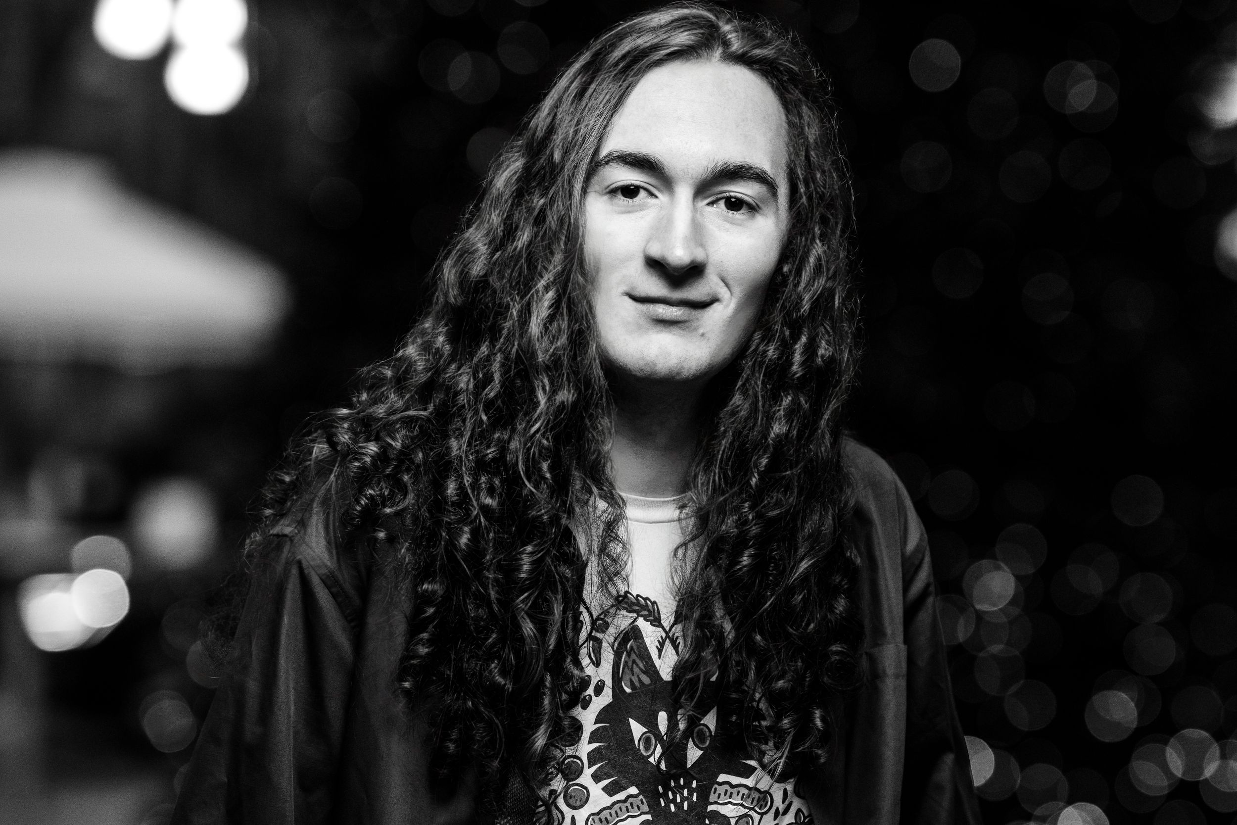 Black and white portrait showing a person with long wavy dark hair smiling while wearing a dark jacket against blurred lights.