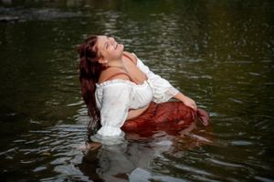 A person in a white top and red skirt reclines dramatically in dark water during an artistic outdoor photoshoot.