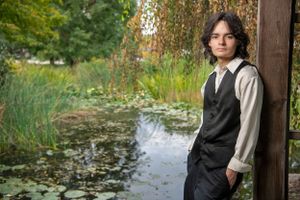Person in formal vest and white shirt leans against wooden post by tranquil pond in garden setting.