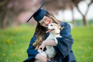 A graduate in blue cap and gown hugs a happy Australian Shepherd dog on a grassy field during graduation photos.