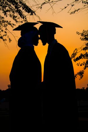 Silhouetted graduates in commencement caps stand close together against a vibrant orange sunset sky framed by tree branches.