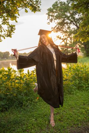 Joyful graduation photo in cap and gown at sunset with arms outstretched.
