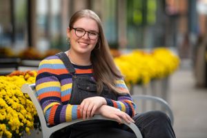 A person in a colorful striped sweater sits smiling on a bench near bright yellow mums in an outdoor setting.