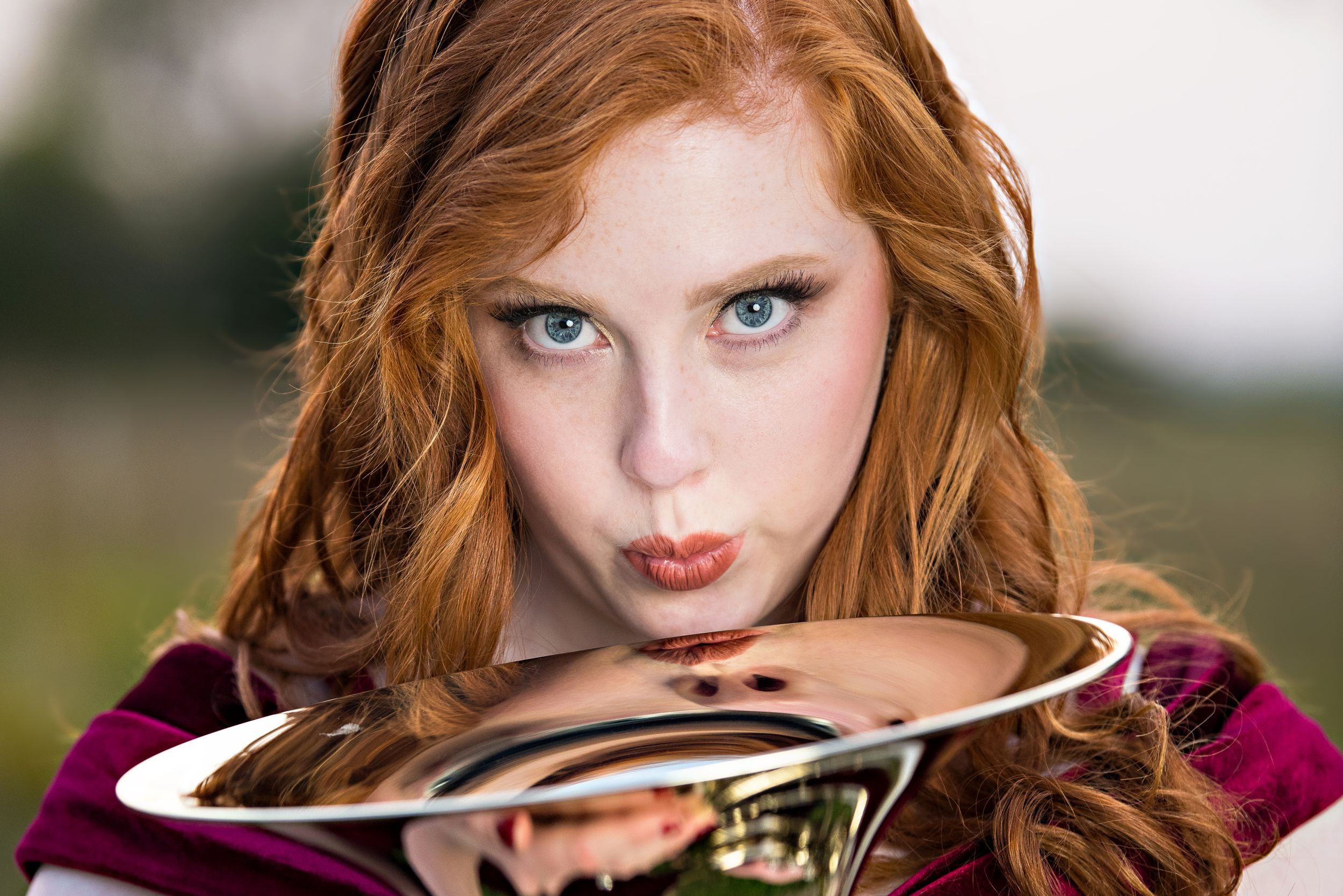 A musician poses with a French horn instrument wearing a burgundy top against a soft focus outdoor background.