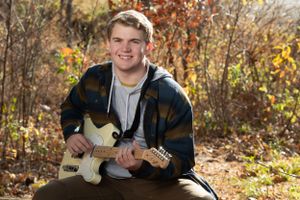 A person playing a white electric guitar outdoors in autumn with colorful fall foliage in the background.