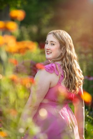A woman in a pink dress poses in a garden filled with vibrant orange and pink flowers on a sunny day.