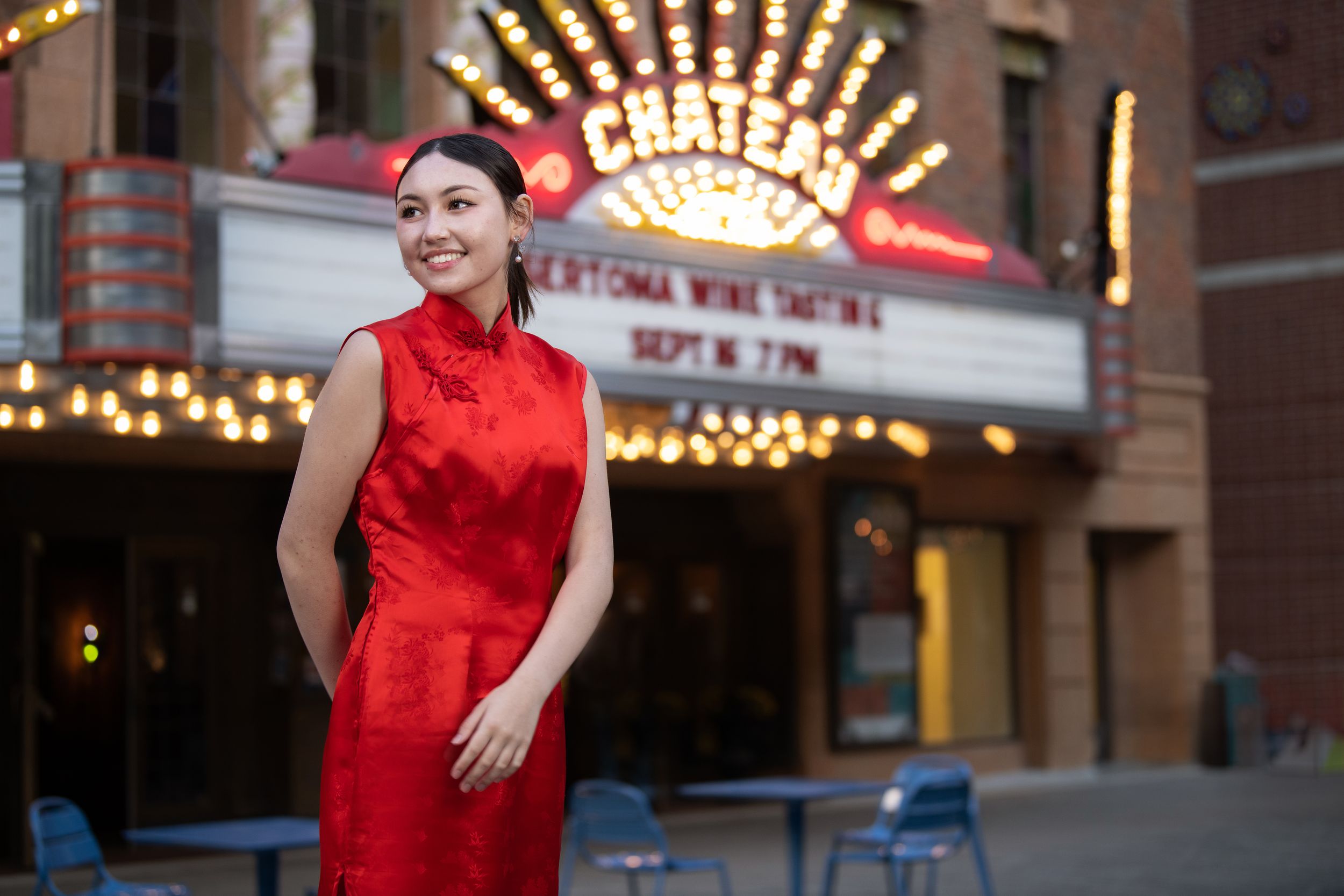 Elegant figure in bright red sleeveless dress poses outside theater with illuminated marquee lights in background.