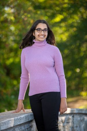 Someone in a pink turtleneck sweater and black pants poses casually on a stone wall outdoors on a sunny day.
