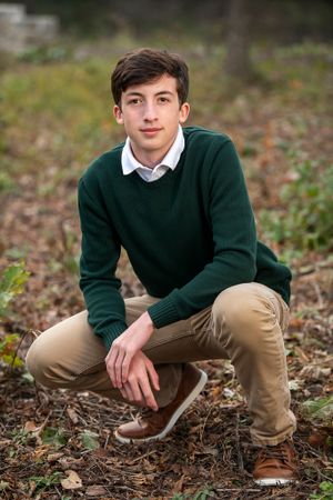 Student in green sweater and khakis crouching outdoors in fallen leaves.